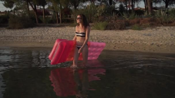 一个长期拍摄的年轻女士走进水中，手里拿着鼓鼓的床垫。女孩走进海里，手里拿着鼓鼓的床垫游泳. — 图库视频影像