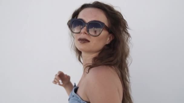 Nahaufnahme einer jungen Frau mit Sonnenbrille, die ihr Haar auf dem weißen Wandhintergrund berührt. — Stockvideo