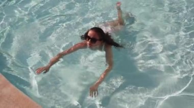 Güneş gözlüklü ve havuzda yüzen beyaz mayo giymiş genç bayanın orta pozu. Güneş gözlüklü ve beyaz mayosu olan genç bir kadın havuzda yüzüyor..