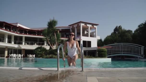 Середній знімок молодої леді в білому вологому купальнику і сонцезахисних окулярах, що виходять з басейну на фоні готелю . — стокове відео