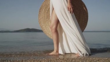 Deniz kıyısında yaz şapkasıyla yürüyen genç bir kadın resmi. Genç bayan, deniz manzarası, bayanların ayakları, su ve şefkat.