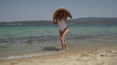 Güneş gözlüklü, yaz şapkalı ve plajda beyaz mayo giyen güzel bir kadın. Genç bayan su kenarında yürüyor ve güneşin tadını çıkarıyor. Güzel, mutlu kadın tatilinin tadını çıkarıyor.