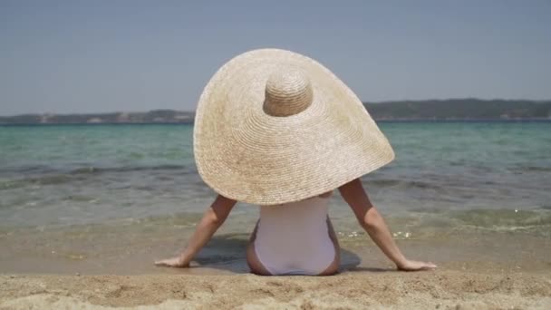 Довгий знімок леді у великому літньому капелюсі і білий купальник на пляжі, милуючись видом. Молода леді сидить біля води і занурюється на сонце. Жінка насолоджується відпусткою на сонці . — стокове відео