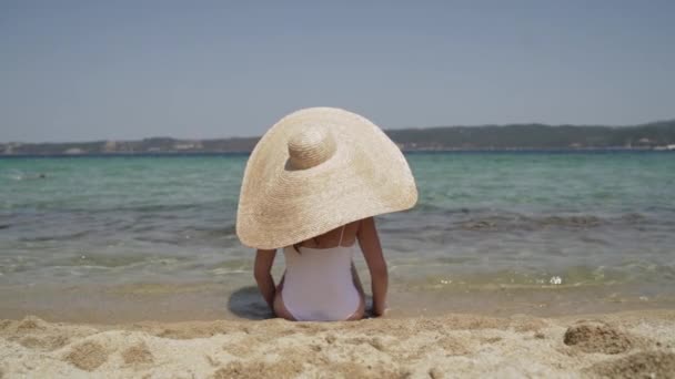 Довгий знімок леді у великому літньому капелюсі і білий купальник на пляжі, милуючись видом. Молода леді сидить біля води і занурюється на сонце. Жінка насолоджується відпусткою на сонці . — стокове відео