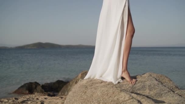 ビーチで長い白いドレスで美しい女性の中間ショット。ビーチの近くの岩で太陽の下で休暇を楽しんでいる素敵な幸せな女性. — ストック動画