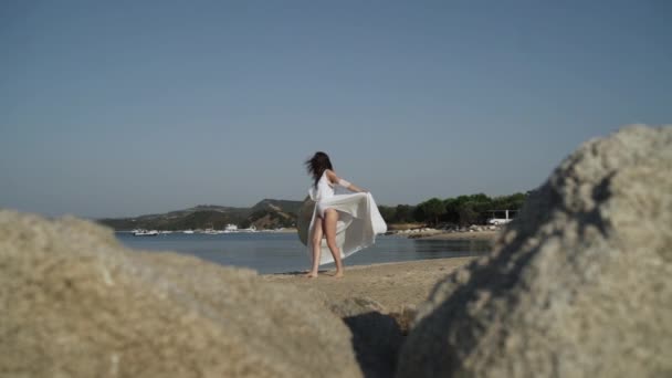 Largo tiro de hermosa dama en vestido blanco girando cerca de la costa. Jovencita con un gran sombrero tomando el sol y disfrutando de sus vacaciones en la playa. Vacaciones de verano y libertad. — Vídeos de Stock