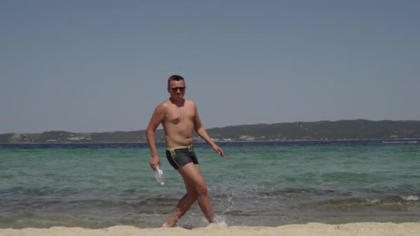 Grote kans dat een man plezier heeft op het strand. Een man in een zonnebril, in een badpak, aan het water aan het trainen. jong mollig man doet workout door de zee. — Stockvideo