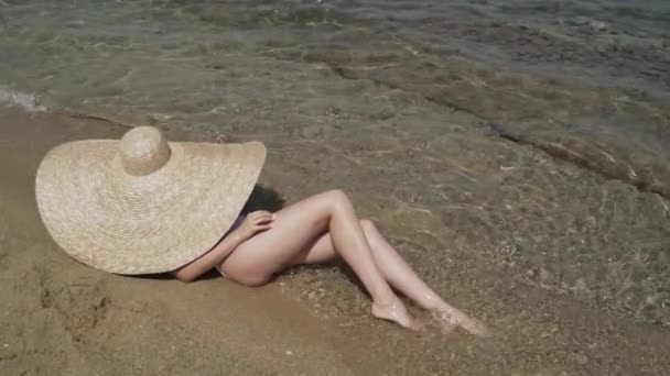 Longue photo de dame en grand chapeau d'été et maillot de bain blanc couché sur la plage dans l'eau. La jeune femme est allongée au bord de l'eau et se prélasse au soleil. Femme profitant de ses vacances au soleil. — Video