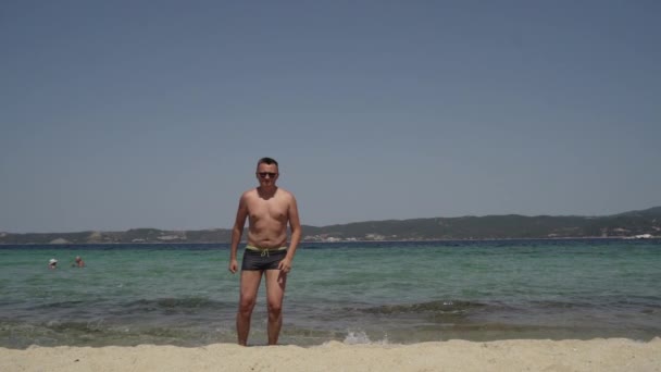 Довгий знімок чоловіка, який розважається на пляжі. Чоловік в сонцезахисних окулярах, в купальнику, вправляється біля води. Молодий пухкий чоловік робить тренування біля моря . — стокове відео