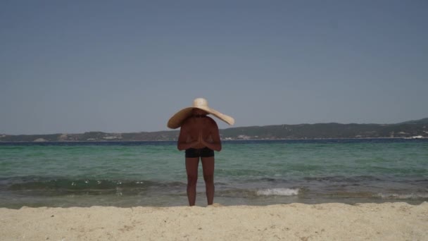 Довгий знімок чоловіка, який розважається на пляжі. Чоловік в сонцезахисних окулярах з великим літнім капелюхом грає біля води, обманюючи навколо і позує . — стокове відео