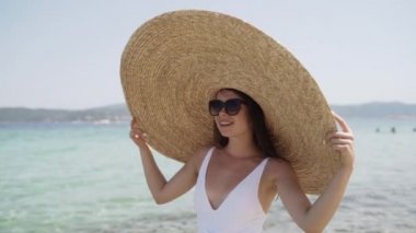 Yazlık şapkalı ve beyaz mayo giymiş, suyun kenarında duran bir kadın resmi. Genç bayan gülümsüyor ve güneşin tadını çıkarıyor. Kadın tatilinin tadını çıkarıyor..