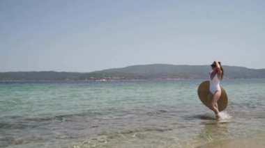 Beyaz mayo giymiş, büyük bir şapkası olan, su kenarında yürüyen ve manzaraya hayran bir kadın. Genç bayan güneşin tadını çıkarıyor. Kadın tatilinin tadını çıkarıyor..