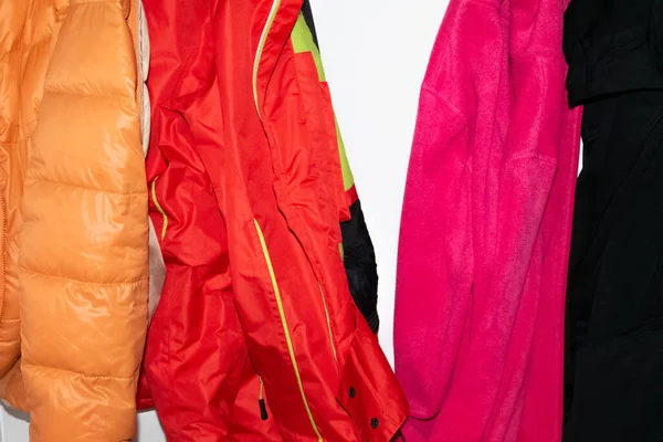 Bright sports orange and red nylon jackets. Soft warm pink and black fleece sweaters, hanging on white wall — Stock Photo, Image