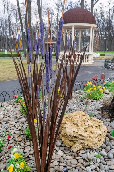 Kharkiv, Ukraine - 17 avril 2021. Installation métallique de roseaux violets et bruns au centre du lit de fleurs, entourée de fleurs de boutons d'or, de grosses et petites pierres. Central Park — Photo