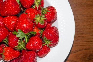 Beyaz bir tabak, ahşap arka planda büyük kırmızı çilek dolu.