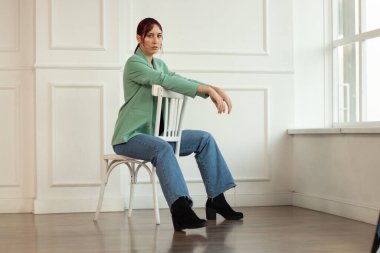 Kızıl saçlı genç bir kızın portresi makyajlı, yeşil ceketli ve kotlu boş bir odada bir koltukta oturuyor.