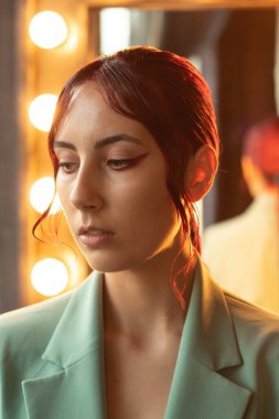 Kırmızı saçlı, makyajlı ve yeşil ceketli bir kızın portresi. Stüdyo lambalarla aynanın karşısında duruyor.