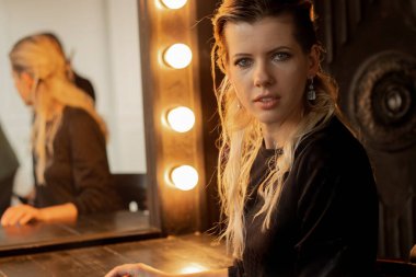 Saçını kestirip akşam makyajı yapan genç bir kız stüdyoda ampullü bir aynanın yanında oturuyor.