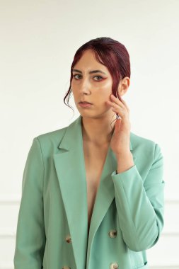 Kırmızı saçlı, makyajlı, yeşil ceketli ve stüdyoda jtnleri olan genç bir kızın portresi.