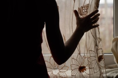 Bir kadının eli yatak odasındaki pencerenin perdesini açıyor, evin penceresinden sokağa bakıyor.