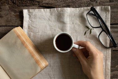 Kadın eli masanın üzerinde açık bir kitap ve bardakların yanında bir fincan kahve tutuyor.