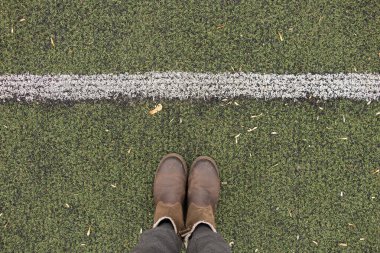 Gri pantolonlu ve kahverengi çizmeli kadın bacakları yeşil çimlerin üzerinde duruyor ve beyaz bir şerit uzanıyor.