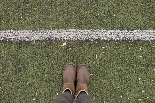 женские ноги в серых брюках и коричневых сапогах стоят на зеленой лужайке и прокладывают разделительную белую полосу