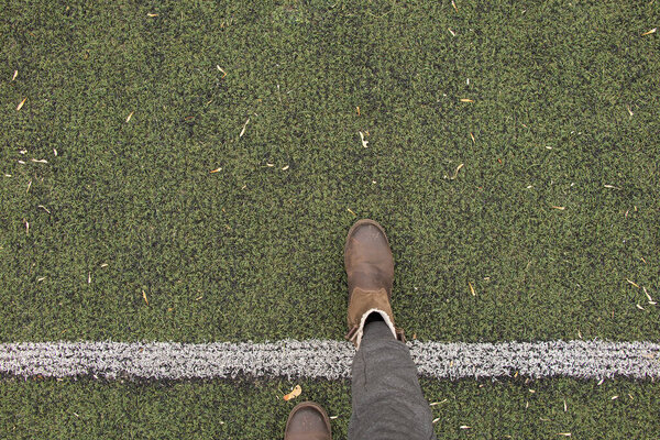 женские ноги в серых брюках и коричневых сапогах стоят на зеленой лужайке и прокладывают разделительную белую полосу