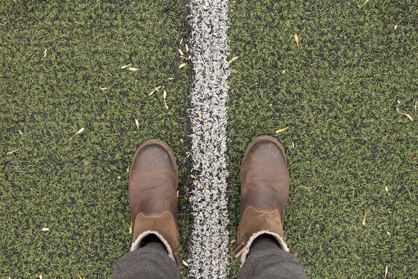 женские ноги в серых брюках и коричневых сапогах стоят на зеленой лужайке и прокладывают разделительную белую полосу