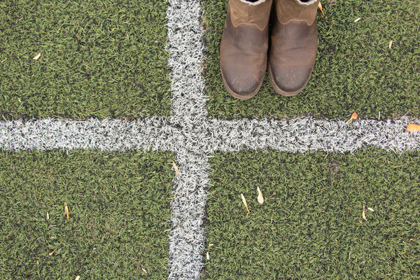 женские ноги в серых брюках и коричневых сапогах стоят на зеленой лужайке и прокладывают разделительную белую полосу
