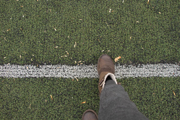 женские ноги в серых брюках и коричневых сапогах стоят на зеленой лужайке и прокладывают разделительную белую полосу