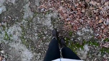 Gri pantolonlu ve kahverengi çizmeli bir kız sonbahar ormanında çimlerin üzerinde yürüyor.