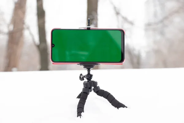 a mobile phone with a green screen in winter stands on a flexible stand in the snow outside