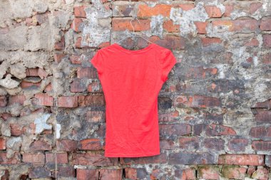 Kırmızı tişört asılıydı. Tuğla duvarda asılı duruyordu. Kadın kıyafetleri giyiyordu.