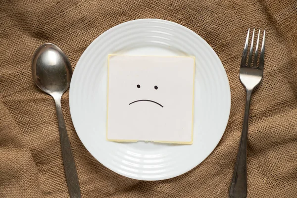 a sad smiley lies on a sheet of paper on a plate next to a spoon and a fork on a brown burlap background, nothing to eat, crisis