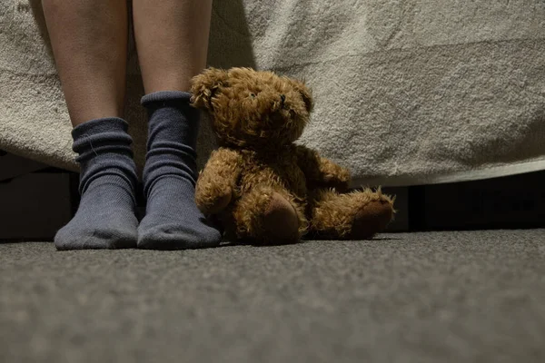 female feet in socks and brown teddy bear on the floor in a dark room