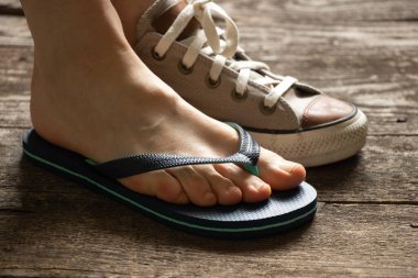 Biri kahverengi tekstil ayakkabılı bir kadın, diğeri de plajda, tahta zeminde parmak arası terlik, tabanında farklı ayakkabılar.