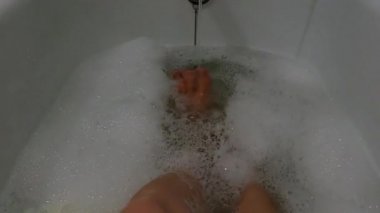 Dişi ayakları sabunlu suyla dolu bir banyoda, bir kız kendini yıkıyor.