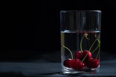 Masadaki siyah arka planda bir bardak suyun içindeki kirazlar, meyve arka planı, meyveler.