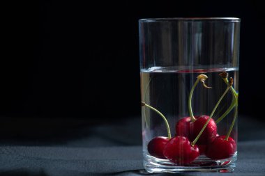 Masadaki siyah arka planda bir bardak suyun içindeki kirazlar, meyve arka planı, meyveler.