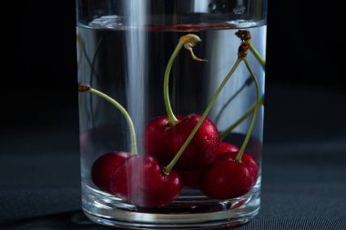 Masadaki siyah arka planda bir bardak suyun içindeki kirazlar, meyve arka planı, meyveler.