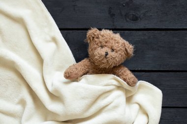 Oyuncak ayı kahverengi ayı beyaz bir battaniyenin altında yatıyor, siyah tahta bir arka planda, çocuk.