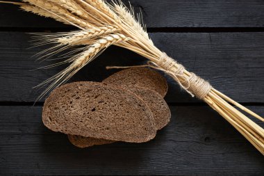 Bir parça siyah ekmek ve siyah tahta bir masanın üzerinde kuru buğday başakları, masada ekmek.