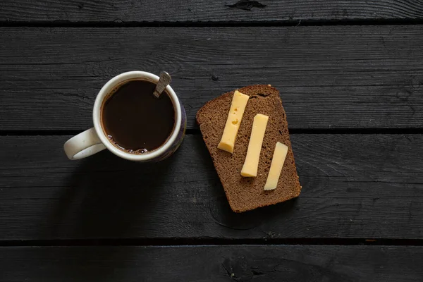 a cup with black coffee and a slice of bread with hard cheese on a black wooden table, homemade coffee for breakfast with a sandwich