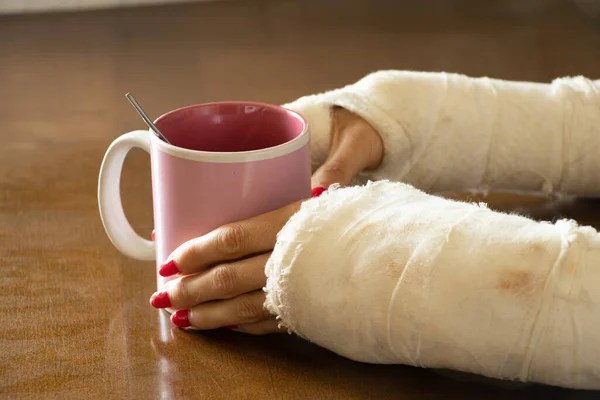 a girl with two broken arms in a cast holds a cup of tea in the kitchen for breakfast, broken arms in a cast, fractured limbs, injury