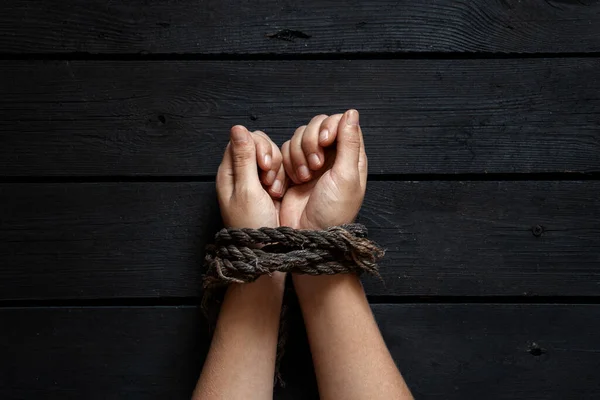 tied female hands with an old rope on a black wooden board background, tied hands, bondage