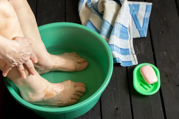 the old grandmother\'s feet are washed in a plastic bowl with water and soap on the black floor, towels were brought, the old grandmother washes her feet in a bowl on the floor, feet were washed