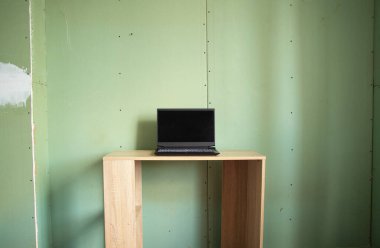 Siyah ekranlı siyah bir dizüstü bilgisayar, duvarları onarılmamış alçı taşı kartondan yapılmış bir evin odasındaki bir masanın üzerinde duruyor, eski bir daire yenileniyor, beyaz ekranlı bir dizüstü bilgisayar.