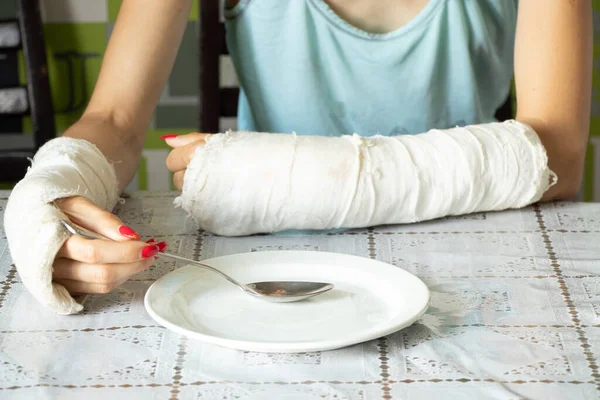 在厨房的饭桌上 一个女孩拿着勺子和盘子一起吃早餐 胳膊被压断了 四肢骨折 — 图库照片