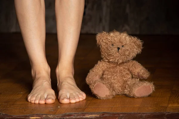 Piedi nudi femminili sollevato un orsacchiotto marrone sul vecchio piano in una stanza buia, giocattolo sul pavimento, camera da letto per bambini — Foto Stock